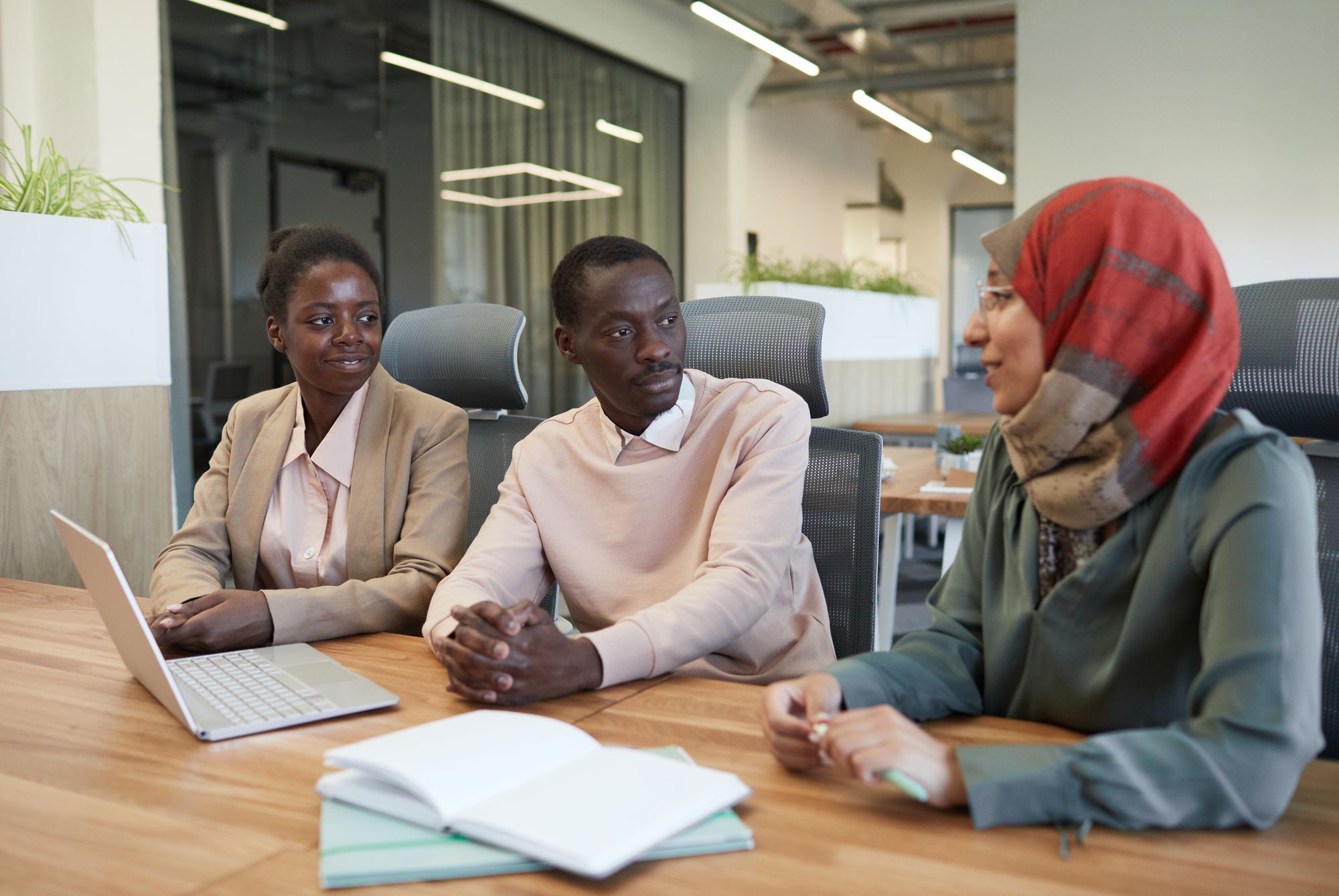 Three professionals in conversation around a wooden table with a laptop and notebooks in a light-filled office.