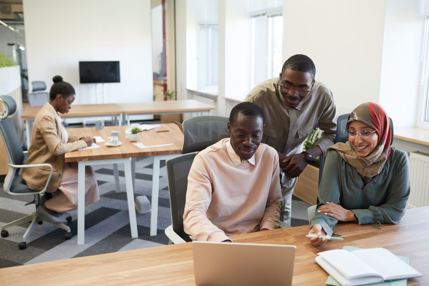 A diverse team of colleagues gathered around a laptop in a bright office, discussing work together.