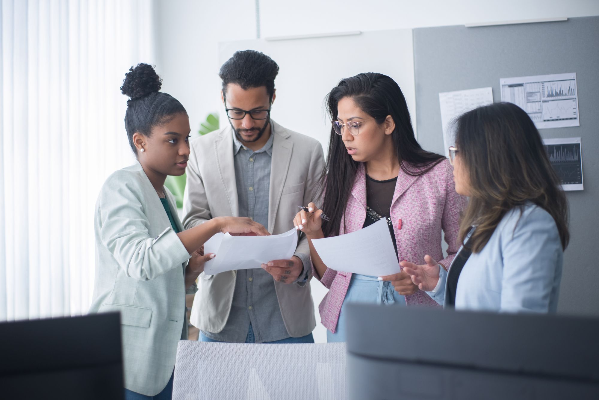 Four colleagues standing together reviewing printed documents, with data charts displayed on the wall behind them.
